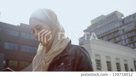 Smiling woman in light beige hijab and blue denim jacket checking phone on sunlit street, buildings Smiling woman in light beige hijab and blue denim jacket checking phone on sunlit street, buildings 139738012