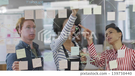 Collaborating team in blue shirt and red-blouse placing sticky notes on glass in office with marker Collaborating team in blue shirt and red-blouse placing sticky notes on glass in office with marker 139738440