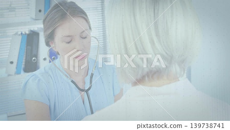 Listening clinician in light-blue blouse auscultating seated patient in exam room with stethoscope 139738741