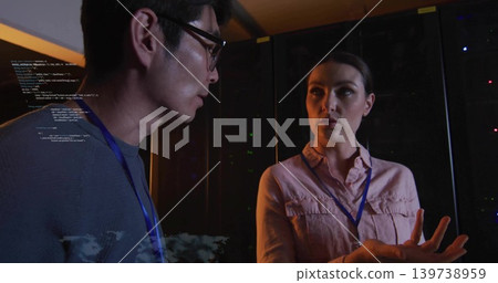 Discussing systems, IT staff gesturing in server room with pink shirt, blue lanyards, code overlay 139738959