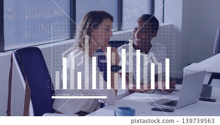 Discussing overlaid bar chart, two women in polka-dot, light blouses using laptop phone blue mug Discussing overlaid bar chart, two women in polka-dot, light blouses using laptop phone blue mug 139739563