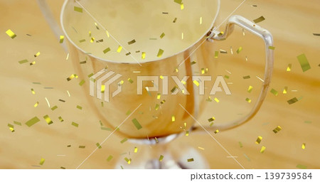 Shining silver trophy cup sitting on light wooden tabletop, gold confetti floating and resting Shining silver trophy cup sitting on light wooden tabletop, gold confetti floating and resting 139739584