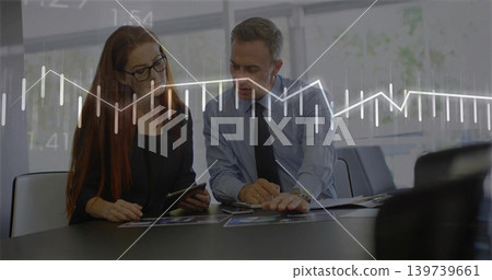 Reviewing woman with tablet while man leaning over charts in office in suits with graph overlay Reviewing woman with tablet while man leaning over charts in office in suits with graph overlay 139739661