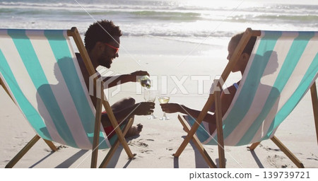 Pouring couple in swimwear seated in striped deck chairs at shore, green bottle and wine glasses Pouring couple in swimwear seated in striped deck chairs at shore, green bottle and wine glasses 139739721