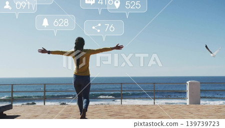 Standing woman in mustard top facing ocean on seafront, with railing, social icons, seagull 139739723