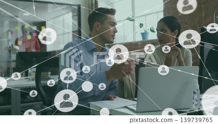Discussing man in blue shirt, woman in sweater examining laptop at desk in office, network overlay Discussing man in blue shirt, woman in sweater examining laptop at desk in office, network overlay 139739765