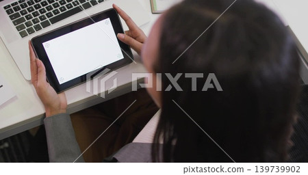 Holding tablet, woman wearing gray blazer viewing blank screen at white desk with laptop and papers 139739902