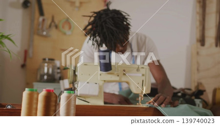 Sitting tailor wearing apron feeding green fabric through machine at workshop, with thread cones Sitting tailor wearing apron feeding green fabric through machine at workshop, with thread cones 139740023
