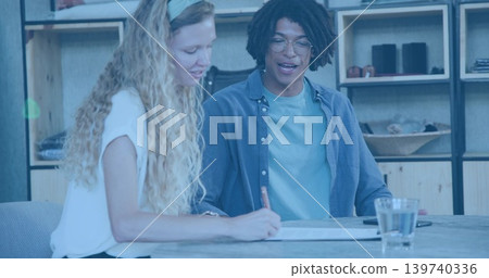 Leaning woman white top headband writing document at wood table, man teal shirt guiding, pen phone 139740336