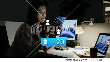 Holding tablet, adult woman wearing light blouse comparing monitors in night cubicle, with alert 20 Holding tablet, adult woman wearing light blouse comparing monitors in night cubicle, with alert 20 139740398
