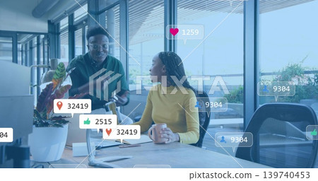 Gesturing pair in green-shirt and mustard-top talking at office desk with laptop mug social-icons Gesturing pair in green-shirt and mustard-top talking at office desk with laptop mug social-icons 139740453