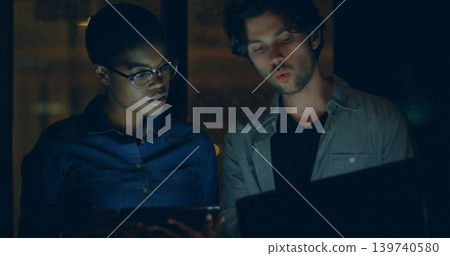 Holding tablet, two colleagues leaning over laptop in dim lit workspace, wearing shirts and glasses Holding tablet, two colleagues leaning over laptop in dim lit workspace, wearing shirts and glasses 139740580