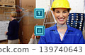 Smiling woman in yellow hard hat, blue coveralls sorting cartons in warehouse with security icons 139740643