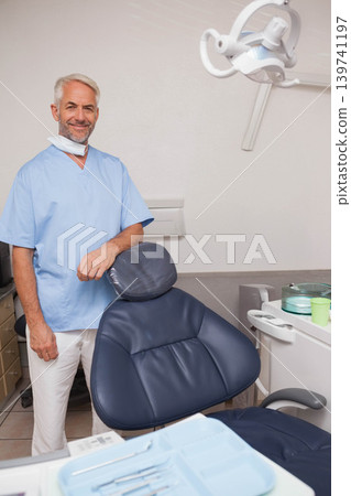 Senior male dentist standing by navy-blue chair in treatment room, wearing scrubs, showing tools Senior male dentist standing by navy-blue chair in treatment room, wearing scrubs, showing tools 139741197