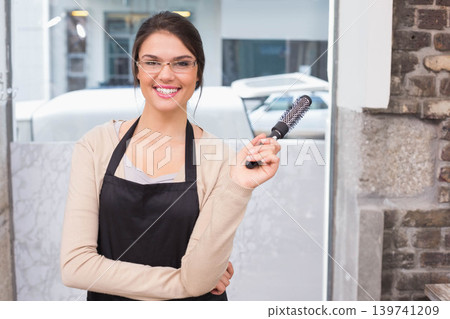 Adult female stylist holding vented brush wearing black apron, clear glasses in salon Adult female stylist holding vented brush wearing black apron, clear glasses in salon 139741209