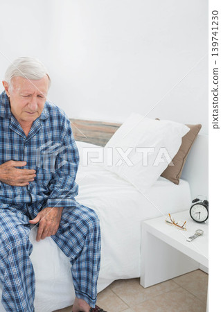 Senior man clutching chest sitting on bed wearing blue plaid pajamas beside alarm clock, copy space Senior man clutching chest sitting on bed wearing blue plaid pajamas beside alarm clock, copy space 139741230