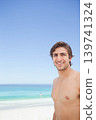 Adult man standing shirtless on sandy beach smiling toward camera with turquoise ocean, copy space 139741324