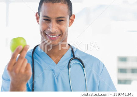 African American male in light blue scrubs holding green apple smiling in clinic with stethoscope 139741343