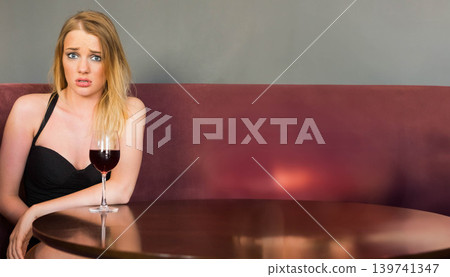 Female sitting in black dress at burgundy booth with stemmed wine glass on table, copy space 139741347