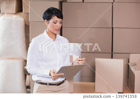 Woman in white shirt and khakis holding clipboard inspecting boxes and bubble wrap in storage 139741358