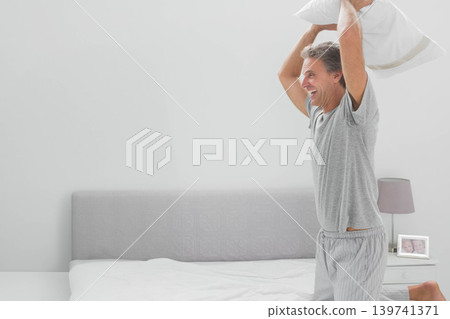 Man wearing gray T-shirt and striped pajama pants holding white pillow overhead on bed, copy space Man wearing gray T-shirt and striped pajama pants holding white pillow overhead on bed, copy space 139741371