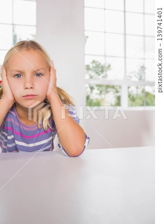 Female child sitting at table pressing hands to ears in striped shirt, grid window, copy space Female child sitting at table pressing hands to ears in striped shirt, grid window, copy space 139741401