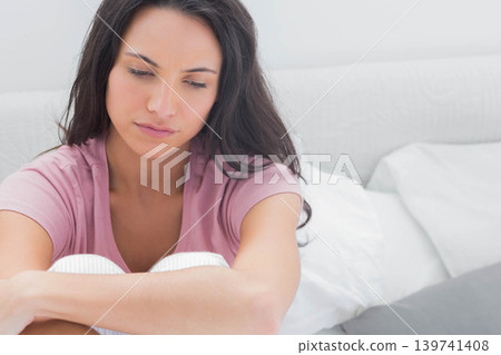 Woman in her twenties sitting on bed wearing pink top, ribbed pants, holding knees among pillows Woman in her twenties sitting on bed wearing pink top, ribbed pants, holding knees among pillows 139741408