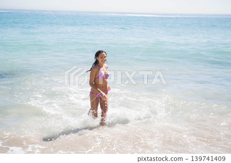 Woman in her twenties wading in shallow ocean at shoreline with patterned swimsuit and foamy surf Woman in her twenties wading in shallow ocean at shoreline with patterned swimsuit and foamy surf 139741409