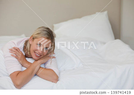 Female lying on bed wearing long-sleeve top with white pillows, propping on elbows, copy space 139741419
