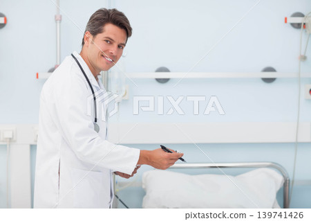Male doctor standing left of ward bed wearing lab coat stethoscope holding chart, copy space Male doctor standing left of ward bed wearing lab coat stethoscope holding chart, copy space 139741426
