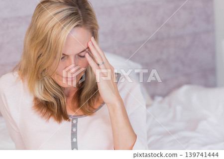 Woman sitting on bed in bedroom wearing long-sleeve top touching temple showing left ring finger Woman sitting on bed in bedroom wearing long-sleeve top touching temple showing left ring finger 139741444