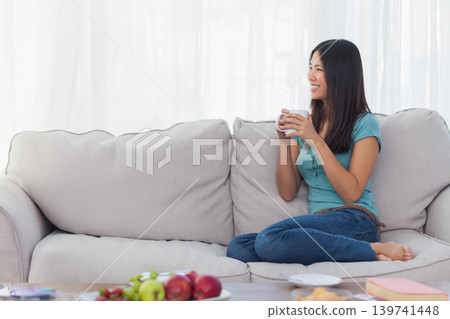 Adult Asian woman sitting cross-legged home holding mug teal top near coffee table, copy space Adult Asian woman sitting cross-legged home holding mug teal top near coffee table, copy space 139741448