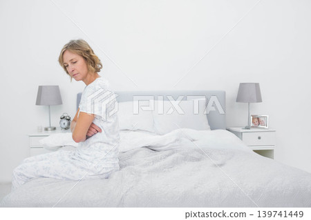 Mature adult female sitting on bed edge, wearing patterned pajamas, beside alarm clock, copy space 139741449