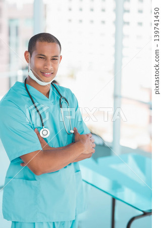 Male clinician standing left near windows in clinic wearing turquoise scrubs, stethoscope and mask Male clinician standing left near windows in clinic wearing turquoise scrubs, stethoscope and mask 139741450