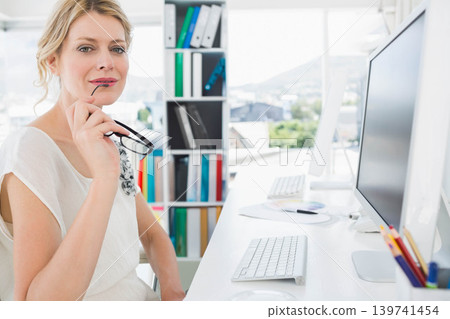 Mature woman sitting at white desk in modern office holding eyeglasses and viewing monitor Mature woman sitting at white desk in modern office holding eyeglasses and viewing monitor 139741454