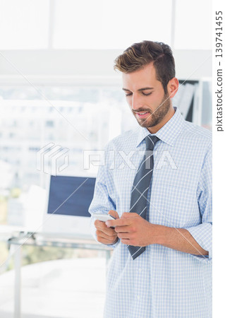Man standing by office window holding phone and checking texts, blue shirt navy tie, copy space Man standing by office window holding phone and checking texts, blue shirt navy tie, copy space 139741455