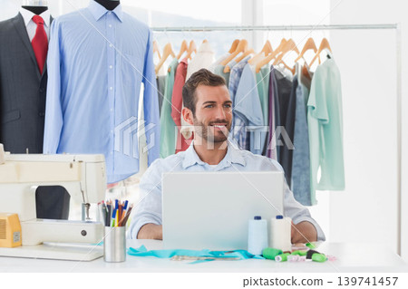 Man working at white table in fashion studio, using laptop and sewing machine near mannequins Man working at white table in fashion studio, using laptop and sewing machine near mannequins 139741457