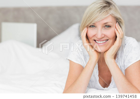 Mature woman reclining on bed, smiling, wearing pajama top, showing ring, bedside book, copy space Mature woman reclining on bed, smiling, wearing pajama top, showing ring, bedside book, copy space 139741458