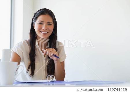 Asian woman sitting at desk in office wearing blouse holding blue pen over open binder, mug Asian woman sitting at desk in office wearing blouse holding blue pen over open binder, mug 139741459