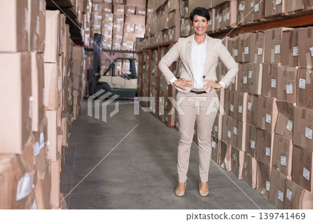 African American mature woman wearing blazer standing hands on hips in warehouse with labeled boxes African American mature woman wearing blazer standing hands on hips in warehouse with labeled boxes 139741469