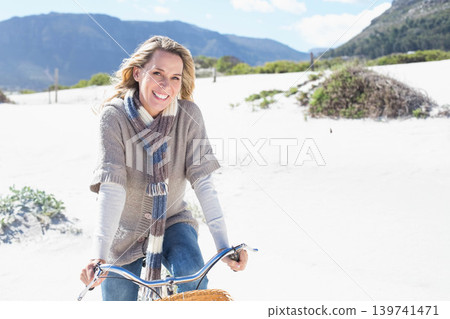 Mature adult woman riding cruiser bicycle across coastal dunes smiling wearing cardigan scarf jeans 139741471