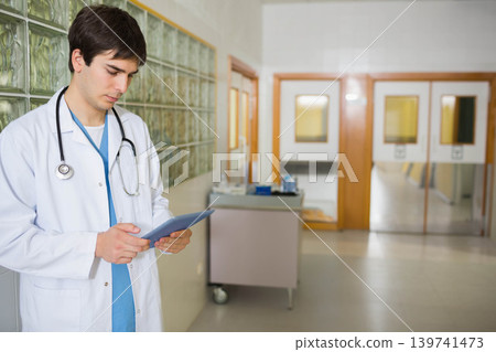 Male doctor checking tablet in hospital corridor, lab coat and scrubs with stethoscope, copy space Male doctor checking tablet in hospital corridor, lab coat and scrubs with stethoscope, copy space 139741473