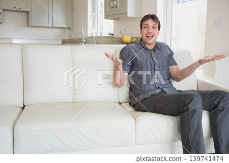 Mid adult man sitting on right white leather sofa shrugging arms in modern home, copy space 139741474