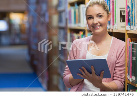 Woman standing and leaning against bookshelf, holding tablet in pink cardigan in library aisle Woman standing and leaning against bookshelf, holding tablet in pink cardigan in library aisle 139741476