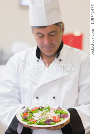Male chef wearing tall white hat and white jacket presenting shrimp salad plate looking in kitchen Male chef wearing tall white hat and white jacket presenting shrimp salad plate looking in kitchen 139741477