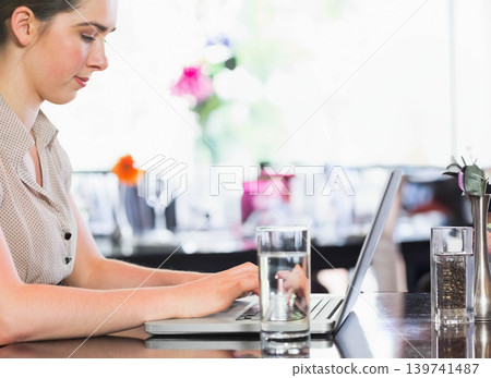 Woman sitting left at reflective cafe table typing on laptop near water glasses, copy space Woman sitting left at reflective cafe table typing on laptop near water glasses, copy space 139741487