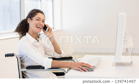 Woman in wheelchair wearing white shirt holding smartphone to ear while using mouse with keyboard 139741497