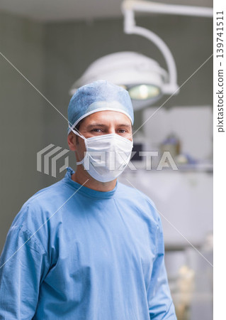 Mid adult male surgeon standing in operating room wearing blue scrubs, cap, mask, overhead lamp Mid adult male surgeon standing in operating room wearing blue scrubs, cap, mask, overhead lamp 139741511