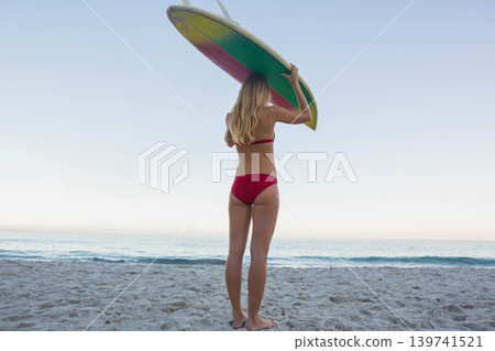 Female in red bikini standing on sandy beach holding colorful surfboard overhead facing sea 139741521