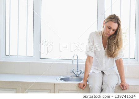 Mid-adult female sitting on countertop near sink, wearing white top, hands resting, copy space Mid-adult female sitting on countertop near sink, wearing white top, hands resting, copy space 139741524